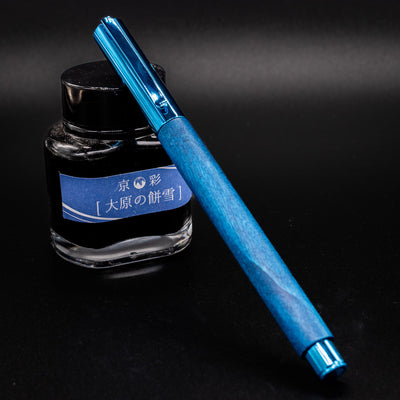 Faber-Castell Ambition Fountain Pen - Blue Wood fully capped, leaning against a Kyo no Oto ink bottle.