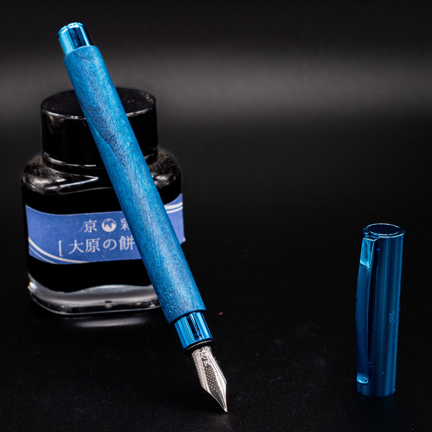 Faber-Castell Ambition Fountain Pen - Blue Wood shown uncapped with nib facing down, resting on a blue-labeled ink bottle.