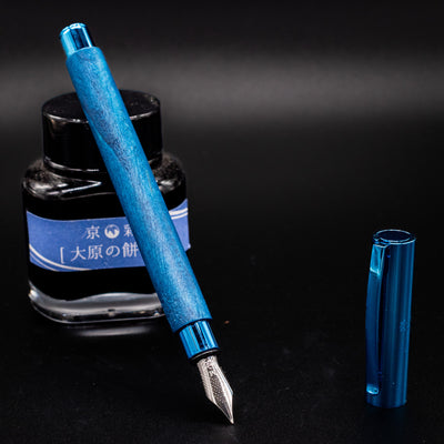 Faber-Castell Ambition Fountain Pen - Blue Wood shown uncapped with nib facing down, resting on a blue-labeled ink bottle.