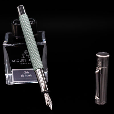 Graf von Faber-Castell Guilloche Fountain Pen in Sage Green with nib exposed, leaning on Jacques Herbin Gris de Houle ink bottle