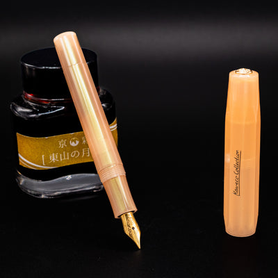 Uncapped Kaweco Collector's Sport Apricot Pearl Fountain Pen beside a gold-labeled ink bottle, revealing gold nib.