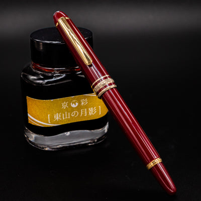 Montblanc Meisterstuck 114R Bordeaux & Gold Mozart Fountain Pen shown capped and resting against a yellow-labeled ink bottle – Preowned