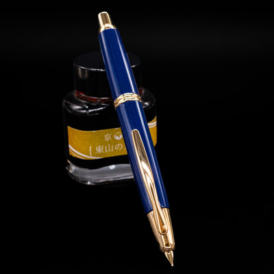 Pilot Vanishing Point Fountain Pen in Blue with Gold Trim – angled view showing nib extended, positioned in front of yellow ink bottle