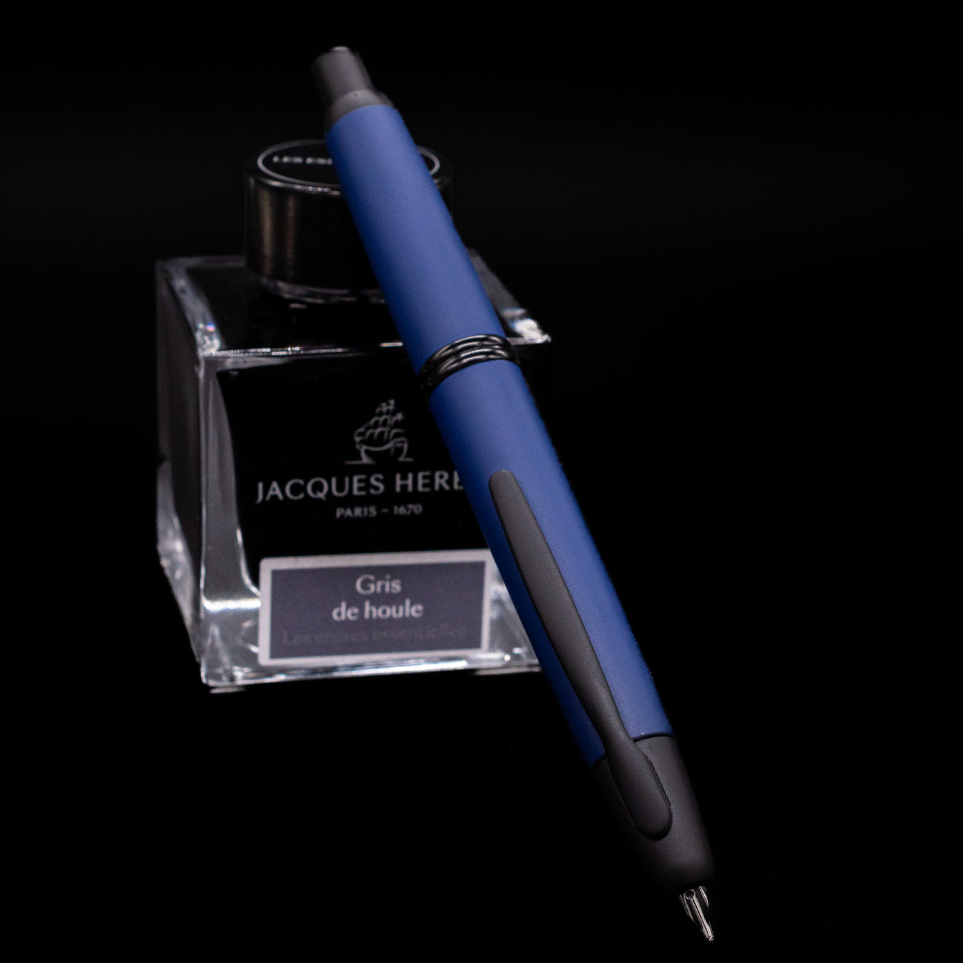 Pilot Vanishing Point Fountain Pen in Matte Blue angled over Jacques Herbin Gris de Houle ink bottle on black background.