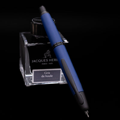 Pilot Vanishing Point Fountain Pen in Matte Blue angled over Jacques Herbin Gris de Houle ink bottle on black background.