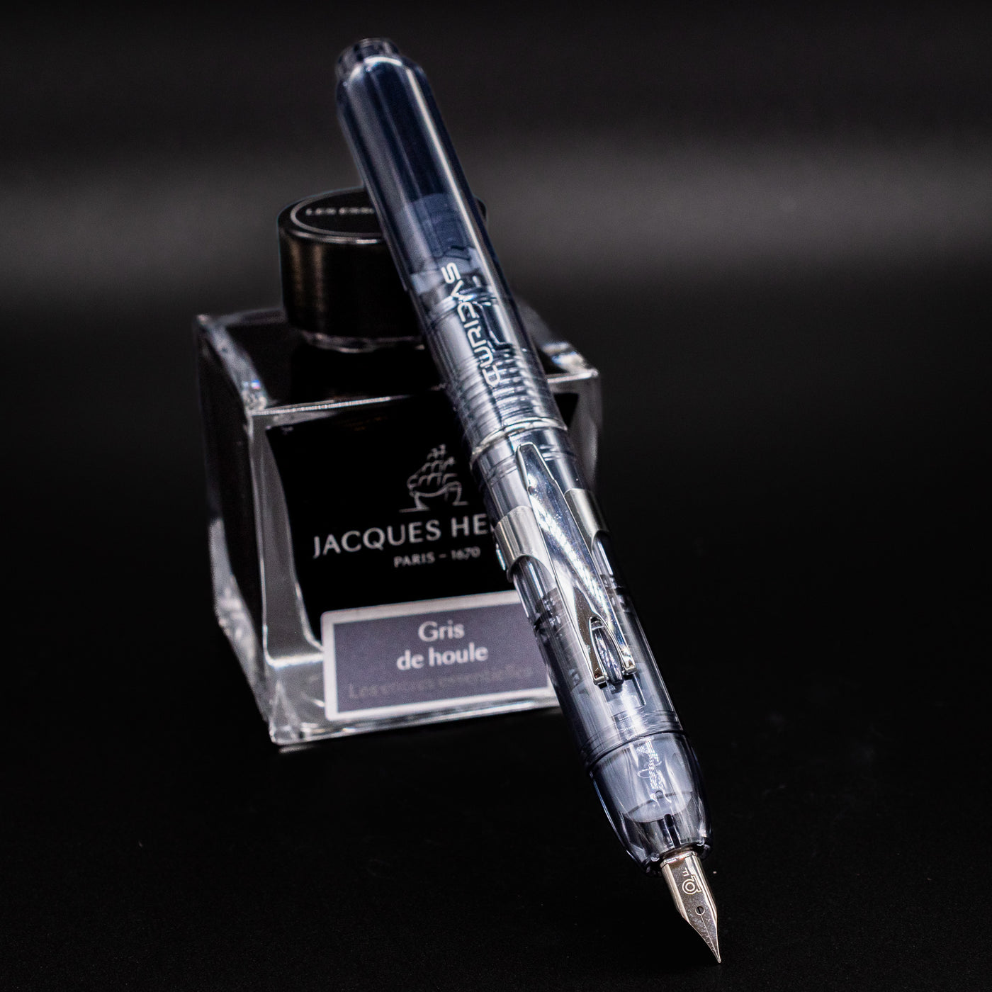 Platinum Curidas Fountain Pen - Graphite Smoke shown in writing position with nib exposed, resting against a bottle of Jacques Herbin Gris de Houle ink.