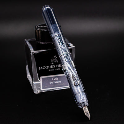 Platinum Curidas Fountain Pen - Graphite Smoke shown in writing position with nib exposed, resting against a bottle of Jacques Herbin Gris de Houle ink.